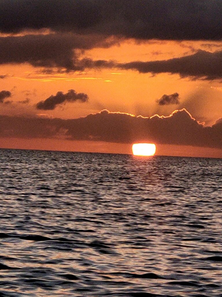"A stunning view of the sun setting over the calm horizon, partially obscured by dark tropical clouds."