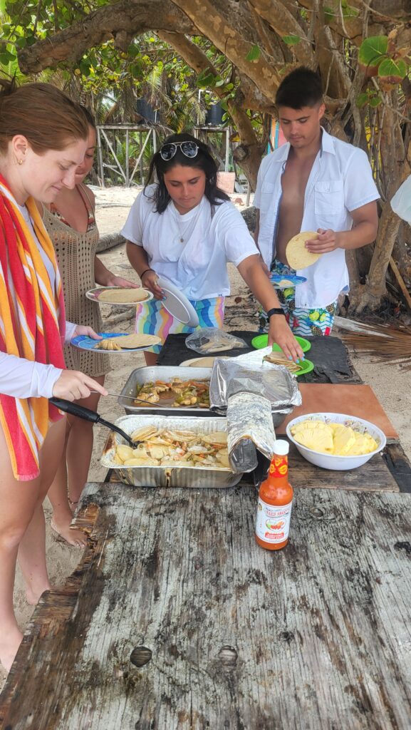 "A group of guests gathered around an outdoor wooden table for a fresh seafood beach cookout in San Pedro."