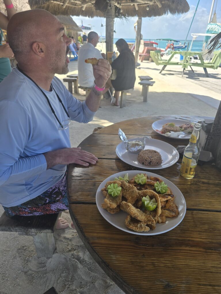 "A guest enjoying a delicious meal of freshly fried fish and local sides at an outdoor beach table."